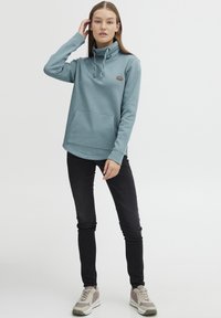 Svijetloplavi pulover s visokim ovratnikom, dodatno teksturiran sitnim točkicama, ima prednju džep i podesivu vezicu. Uparen s crnim uskim trapericama i bež tenisicama.