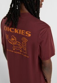Vínová tričko s krátkým rukávem vyrobené z bavlny, s oranžovou grafikou kreslené postavy a textem "DICKIES ON THE ROAD AGAIN."