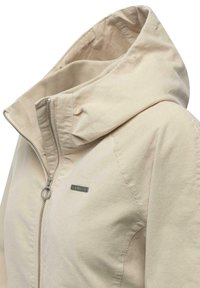 Lyse beige jakke med hætte, der har en lynlås foran og et lille metallogo på brystet. Tekstureret stof med syningsdetaljer.