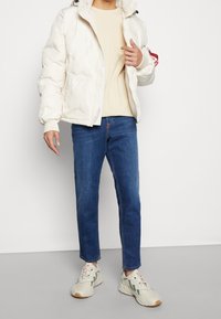 Weiße Daunenjacke mit Reißverschluss und Kapuze, beiges Sweatshirt, blaue Jeans und hellfarbige Sneaker, die einen lässigen Lagenlook präsentieren.