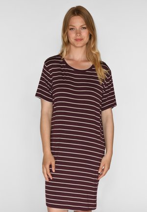 Femme portant une robe courte à manches courtes, longueur genou, avec des rayures blanches horizontales sur un fond marron foncé, debout devant un fond uni.