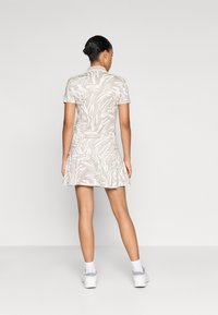 Beiges, kurzärmliges Kleid mit abstrakten weißen Mustern, ausgestelltem Rock und Kragen. Das Material wirkt glatt und leicht. Getragen mit weißen Sneakers.