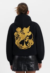 Zwarte hoodie met een capuchon, voorzien van een opvallend gouden bloemenborduurontwerp op de achterkant. Het materiaal lijkt zacht, met een ontspannen pasvorm.
