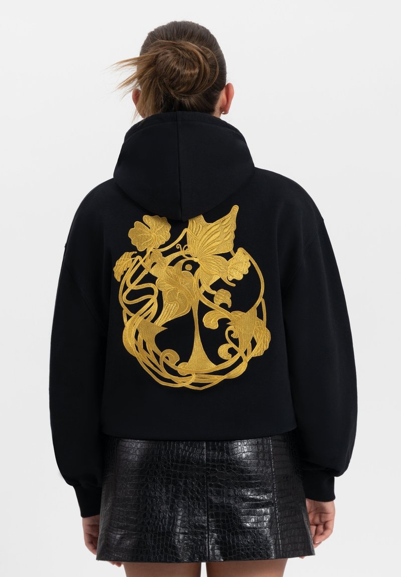 Zwarte hoodie met een capuchon, voorzien van een opvallend gouden bloemenborduurontwerp op de achterkant. Het materiaal lijkt zacht, met een ontspannen pasvorm.