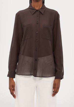 Semigennemsigtig mørkebrun skjorte med lange ærmer og knapper samt to brystlommer, båret med hvide bukser.