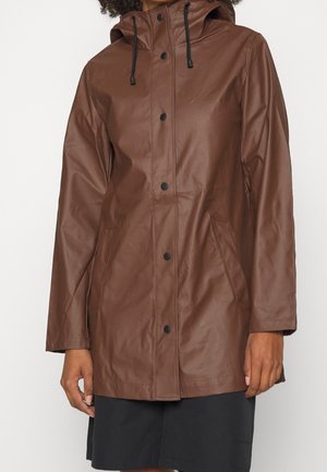 Femme portant un imperméable à capuche marron avec des boutons-pression noirs et des poches latérales, assorti à une jupe noire.