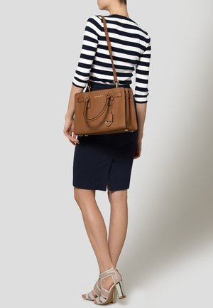 Hnědá kožená kabelka s dvojitými horními uchy, zlatými kovovými doplňky a strukturovaným designem, nošená přes pruhovaný top a tmavě modrou sukni.