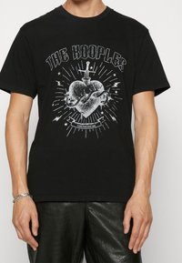 Camiseta de algodón negra con un estampado gráfico de un corazón con una espada y una serpiente, superpuesto con rayos y el texto "THE KOOPLES." Corte ajustado.