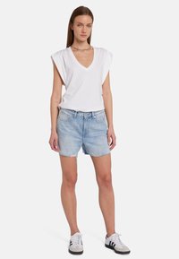 T-shirt bianca con scollatura a V e pieghe sulle spalle, abbinata a pantaloni corti in denim azzurro chiaro con orli sfrangiati, indossati con sneakers bianche e nere.