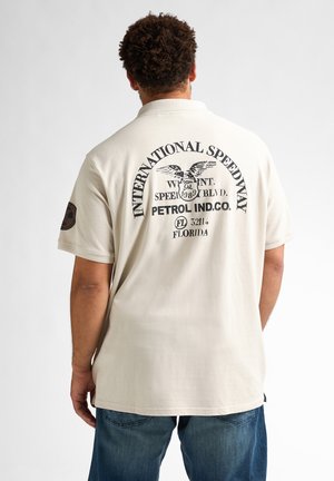 Creme kleurige polo shirt met korte mouwen, voorzien van een grote zwarte print op de achterkant met een adelaar en tekst. De stof lijkt glad en casual.