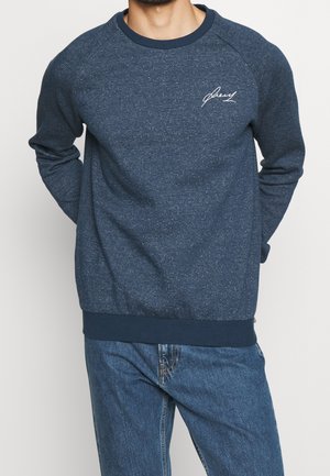 Mand iført marineblå tekstureret sweatshirt med hvidt skriftslogo på brystet og blå jeans, hænderne bag ryggen.