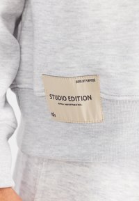 Svijetlosivi dukserica s teksturiranim materijalom, rebrastim rukavima i bež oznakom s natpisima "STUDIO EDITION" i "ALPHA INDUSTRIES INC."