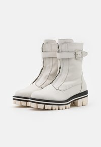 Botines de cuero blanco con una correa de hebilla, que cuentan con una suela gruesa beige con acentos negros y detalles de costura a lo largo de las costuras.