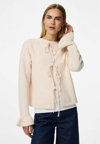 Mujer rubia con cardigan de punto crema con lazos y puños acampanados sobre una camisa blanca, combinando con vaqueros azul oscuro, posando con la mano cerca de la oreja.