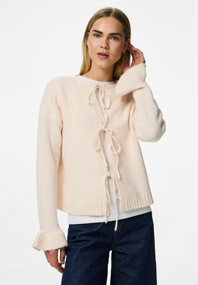 Mujer rubia con cardigan de punto crema con lazos y puños acampanados sobre una camisa blanca, combinando con vaqueros azul oscuro, posando con la mano cerca de la oreja.