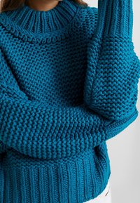 Pull tricoté turquoise avec col côtelé. Présente une texture épaisse, une coupe oversize et des accents texturés sur les manches.