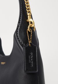 Sac à main en cuir noir avec une texture lisse, doté d'une anse en chaîne au ton doré, d'un détail de tag et d'accents de couture. Marque Coach visible.
