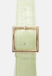 Vaaleanvihreä vyö, jossa on kohokuvioitu krokotiilimalli, varustettuna kultaisella neliskulmaisella soljella ja valkoisella ompeleella reunoilla.