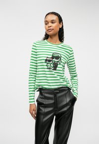 Zöld és fehér csíkos, hosszú ujjú póló kereknyakú szabással, melyen két karakter grafikai nyomata látható. Fekete műbőr nadrággal párosítva.