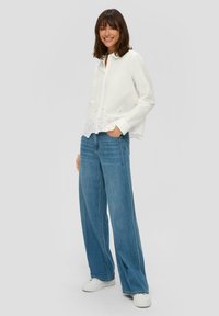 Weißes Hemd mit Knopfleiste und Lochspitze am Saum, kombiniert mit hellblauen, weiten Jeans und weißen Sneakers.