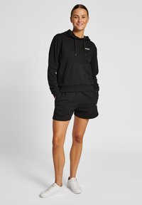 Svart hoodie med framficka och snörning på huvan, ihop med svarta shorts. Skorna är vita sneakers. Enkel, avslappnad design.