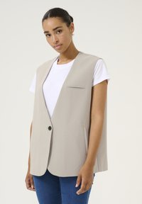 Beige sleeveless vest with a single button, a chest pocket, and side pockets, made of smooth fabric. Worn over a white t-shirt.