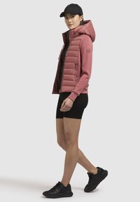 Gevoerde roze jas met een capuchon, voorzien van een rits aan de voorkant en zijzakken, gedragen over een zwart top en shorts, gecombineerd met zwarte sneakers.