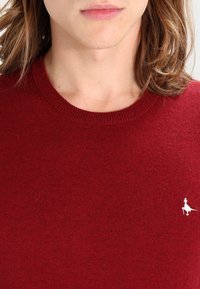 Rode gebreide trui met een ronde hals en geribde afwerking. Bevat een klein wit logo van een figuur op de linkerkant van de borst. Zachte textuur.