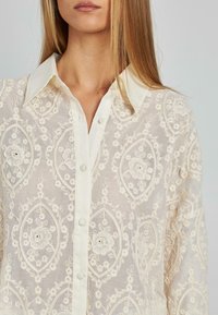 Bestickte Bluse aus einem transparenten, leichten Stoff, mit Blumenmustern, einem spitzen Kragen und Knopfverschlüssen auf der Vorderseite.