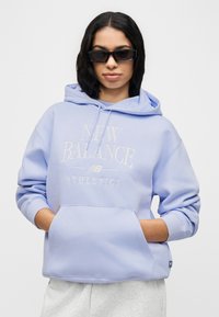 Νεαρή γυναίκα με μαύρα γυαλιά ηλίου, ελαφρύ μωβ φούτερ New Balance και ανοιχτό γκρι παντελόνι, με τα χέρια στην τσέπη του φούτερ, μπροστά σε λευκό φόντο.