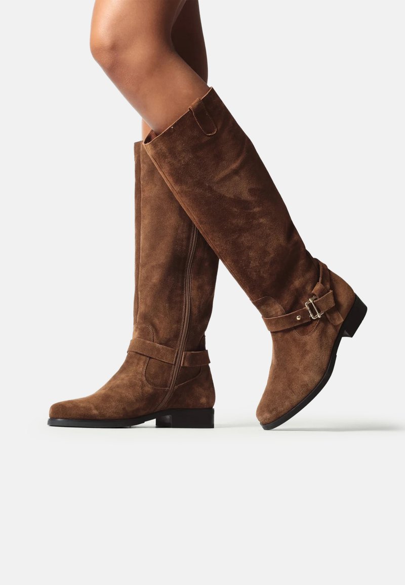 Bocanci din nubuc maron înalt până la genunchi, cu talpă plată, având un fermoar lateral, accent de cataramă și o textură netedă pentru confort și stil.