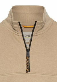 Beige kwart-rits trui van zacht materiaal, met een zwarte rits met een getextureerde trekker en een label met "camel active" in de kraag.