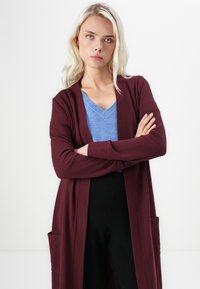 Bordeaux lange cardigan met een open voorkant en zakken, gelaagd over een blauwe V-hals trui. Model staat met gekruiste armen voor een witte achtergrond.