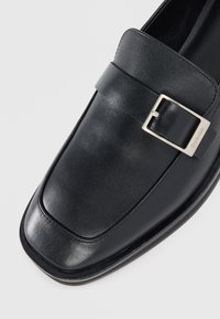 Mocassino in pelle nera con punta quadrata e dettaglio di fibbia argentata sulla cinghia, con un sottile marchio Calvin Klein sul bordo della fibbia.