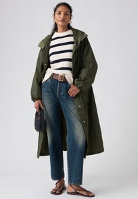 Long manteau vert porté sur un pull rayé blanc et marine, jean bleu avec une finition délavée, et sandales marron. Elle tient une petite pochette noire.