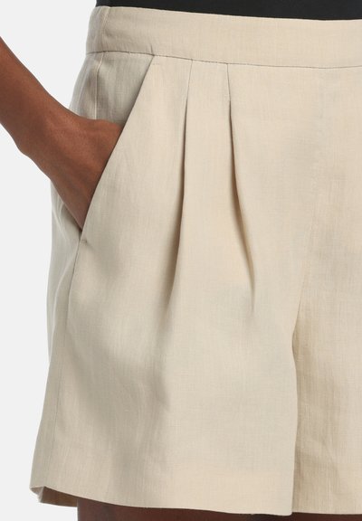 Nærbillede af beige linnedshorts med høj talje, forlommer med læg og en hånd i venstre lomme.