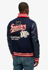Chaqueta varsity de color navy con cuello a rayas rojas y blancas. Presenta logos bordados, un gráfico de un tigre y mangas de cuero. Se muestra la vista trasera.