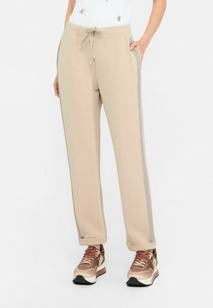 Beżowe spodnie dresowe z błyszczącymi paskami po bokach, elastyczna talia ze sznurkiem, w połączeniu z brązowymi i beżowymi trampkami, ręce w kieszeniach.