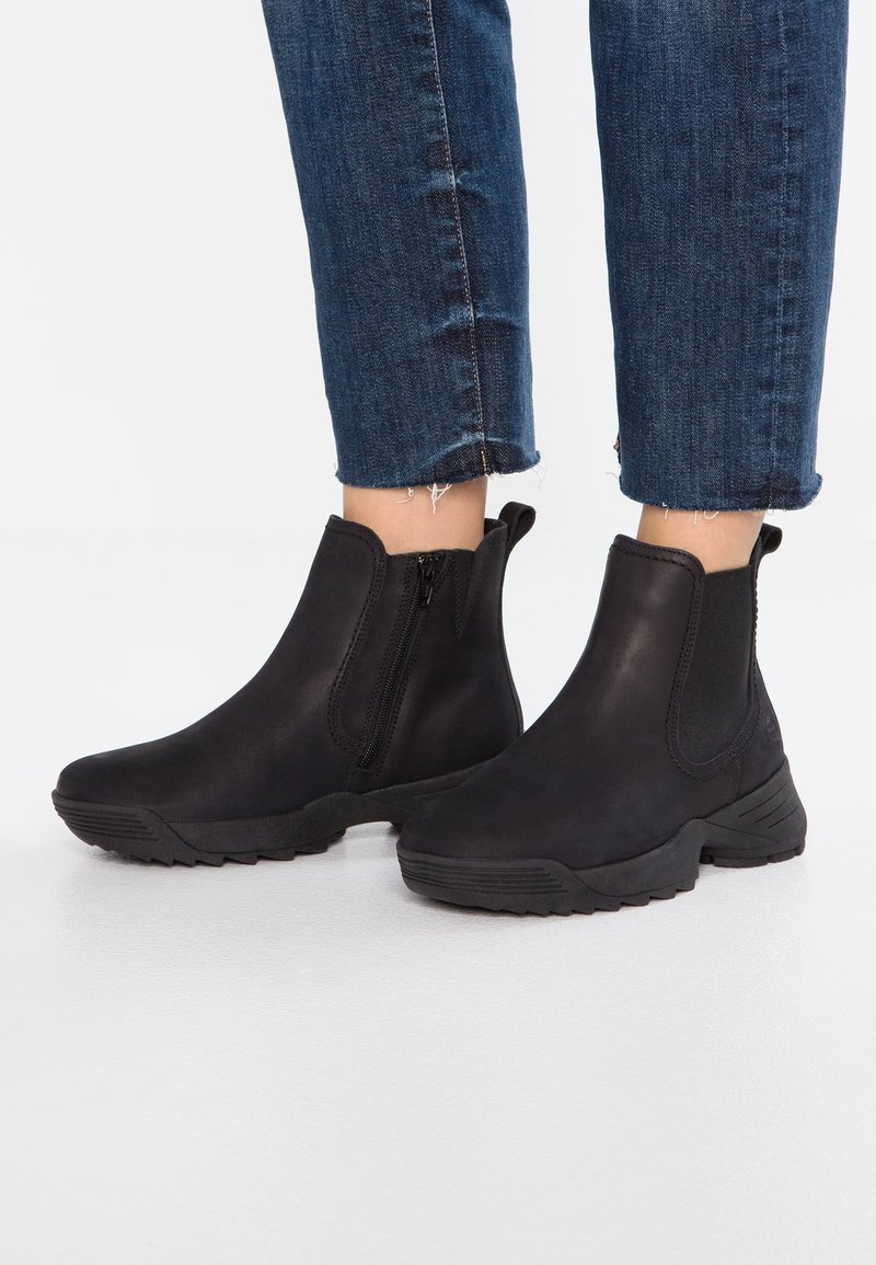 Bottes noires à cheville avec fermeture éclair latérale et panneaux élastiques portées avec un jean bleu coupé sur fond blanc.