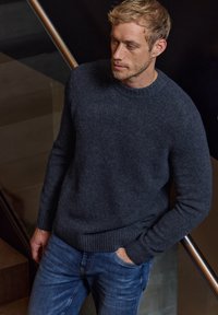 Dunkelgrauer Wollpullover mit Rundhalsausschnitt, gerippten Bündchen und Saum, kombiniert mit blauen Jeans. Der Hintergrund zeigt eine Treppe.