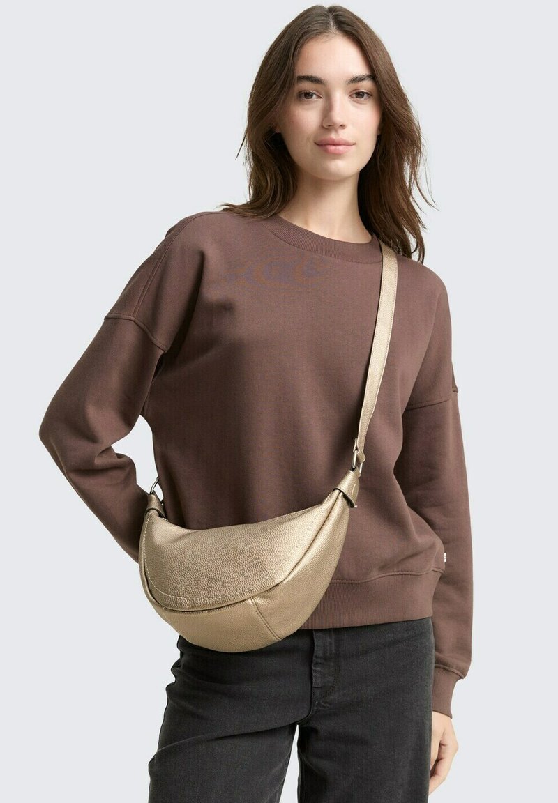 Borsa a tracolla dorata, testurizzata, con forma curva e manico; indossata con una felpa marrone e jeans neri. Design semplice, pochi dettagli.