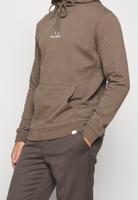 Persona que lleva una sudadera con capucha marrón con el logo "LES DEUX" y pantalones marrones a juego, con una mano en el bolsillo.