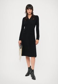 Sieviete valkā melnu garu piedurkņu adītu kleitu ar divām priekšējām kabatām, tur rokā melnu somiņu ar zelta ķēdēm un uzvelk melnus potīšu zābakus.