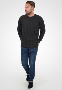 Jersey de punto gris oscuro con textura acanalada, cuello redondo y mangas largas, combinado con jeans azules y zapatos negros.