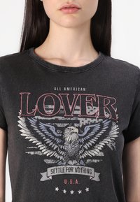 T-paita, jossa on hiilenharmaa puuvilla, ja siinä on graafinen kuva, jossa on kotka, Yhdysvaltain lippu ja teksti "ALL AMERICAN LOVER" paksulla punaisella ja harmaalla värillä.