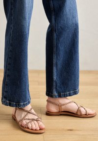 Sandales plates tan avec des lanières tressées et un design minimaliste, associées à un jean large bleu, sur un fond de sol en bois.