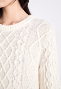 Femme portant un pull en maille torsadée de couleur crème avec des motifs en losange et tressés, montré de l'épaule au cou.