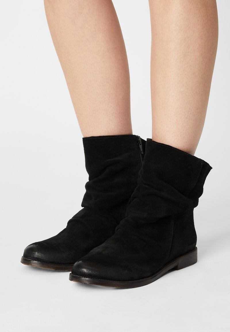 Jambes inférieures portant des bottes en daim noir souples mi-mollet avec des bouts ronds et des fermetures éclair latérales, debout sur une surface blanche.