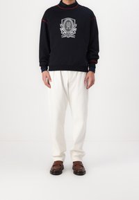 Sudadera marina con acentos rojos y un logo gráfico, combinada con pantalones blancos holgados y zapatos de lona marrón. Conjunto casual.