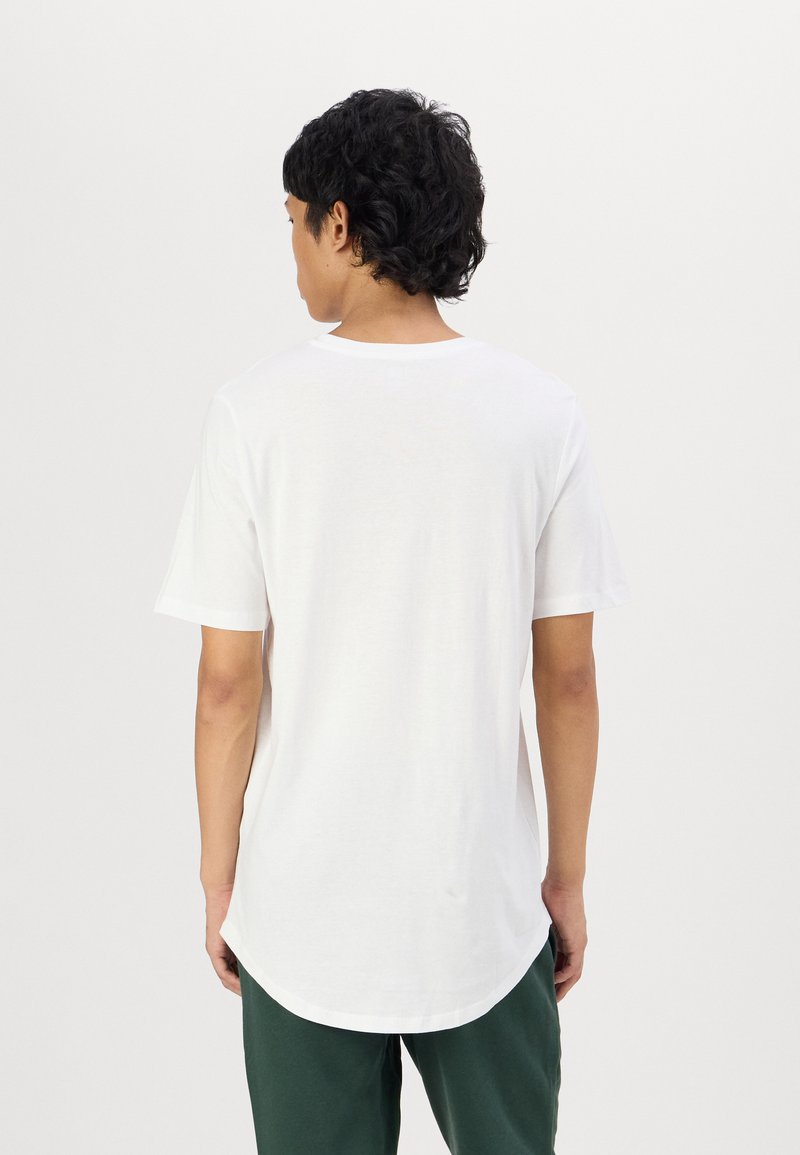 Weißes T-Shirt mit kurzen Ärmeln und rundem Halsausschnitt, mit einem längeren hinteren Saum, aus weichem Stoff, getragen mit grüner Hose.